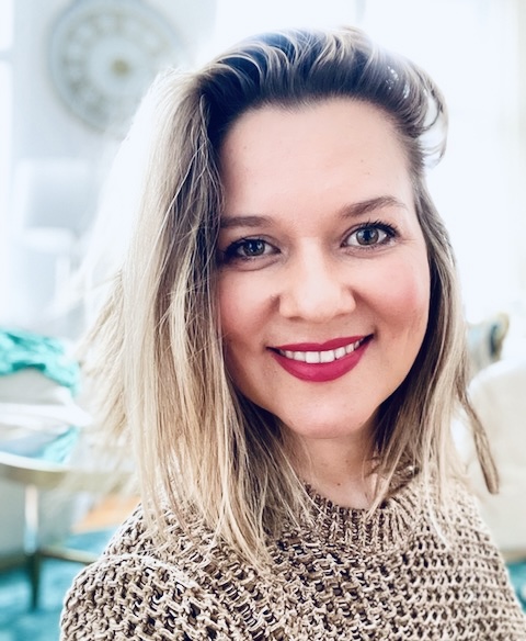 A fair-skinned woman with blonde hair and a beige knit sweater smiles at the camera, set against a blurred background feat...