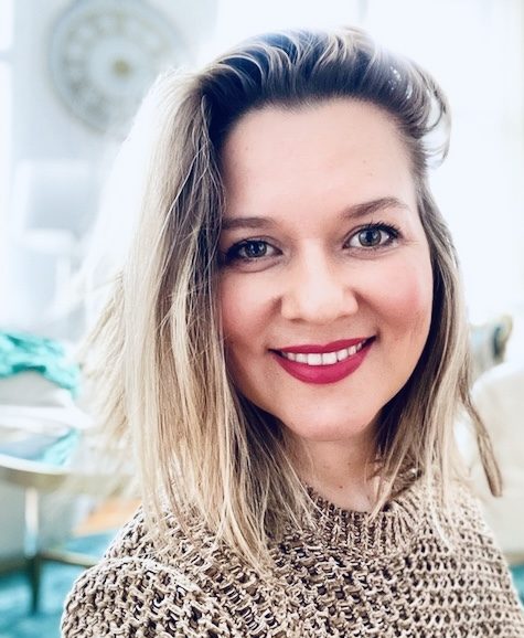 A fair-skinned woman with blonde hair and a beige knit sweater smiles at the camera, set against a blurred background feat...
